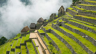 The ancient stone ruins and green agricultural terraces of the Incan citadel Machu Picchu are built into a mountainside, with clouds in the background.