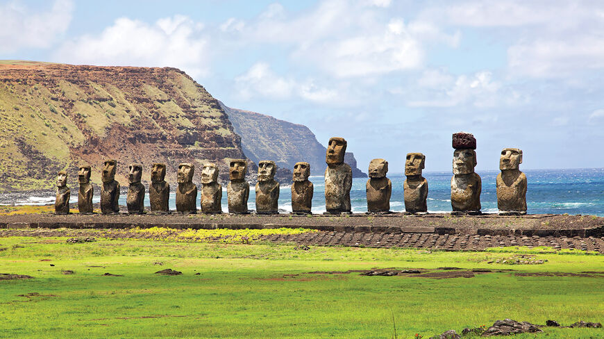 Fifteen stone Moai statues stand on a platform in Rapa National Park on Easter Island, with a backdrop of the ocean and rugged cliffs.