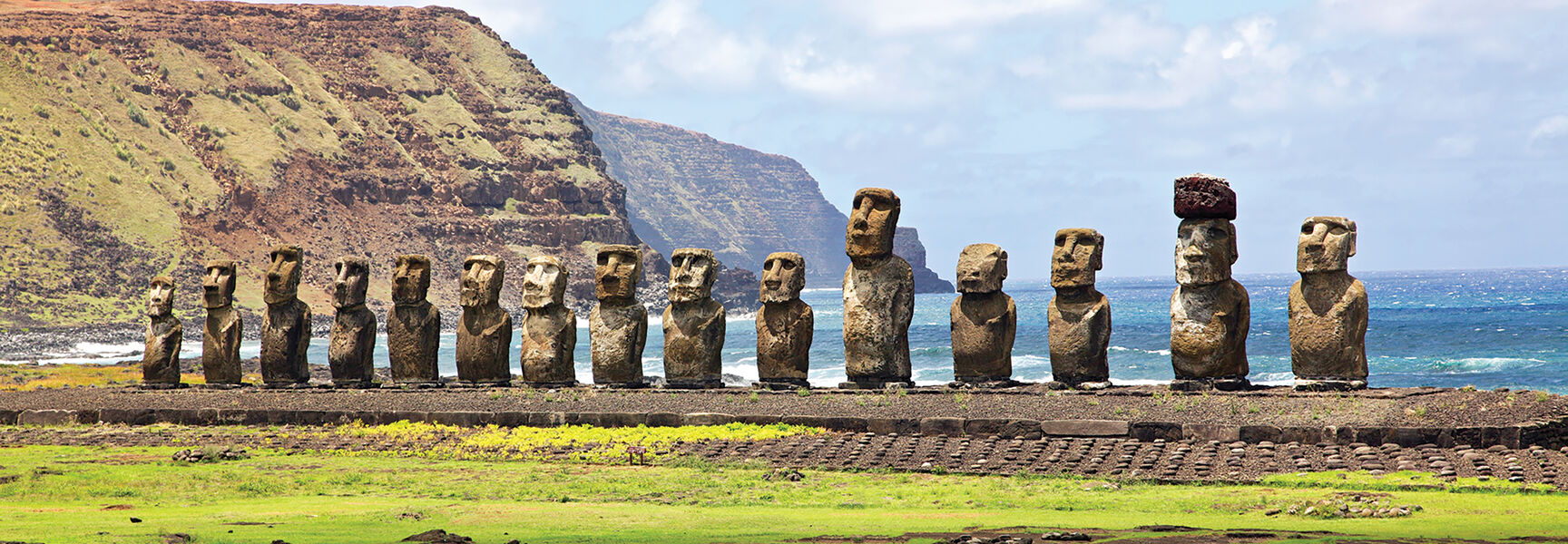 Fifteen stone Moai statues stand on a platform in Rapa National Park on Easter Island, with a backdrop of the ocean and rugged cliffs.