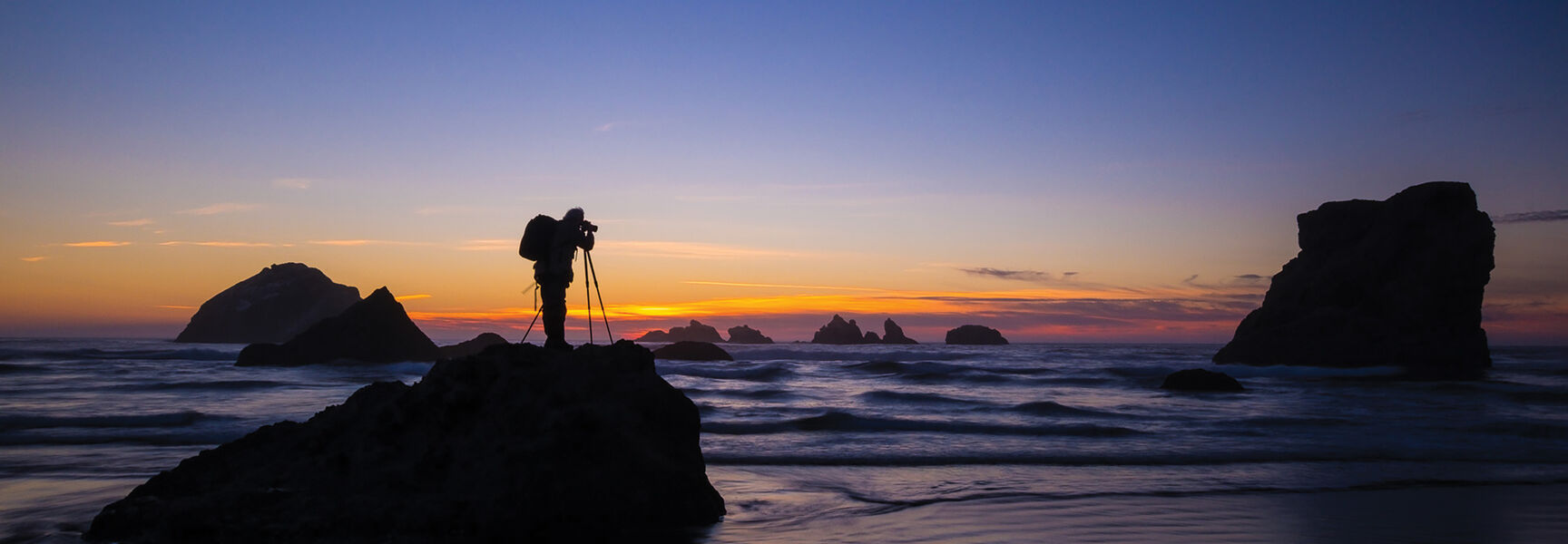 Silhouette of a photographer capturing a sunset landscape on a rocky beach, illustrating advanced digital photography techniques.