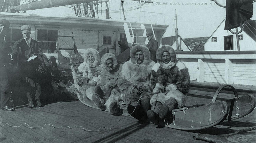 Four men from Peary's North Pole Expedition wearing heavy fur parkas sit on a wooden sled on the deck of a ship.