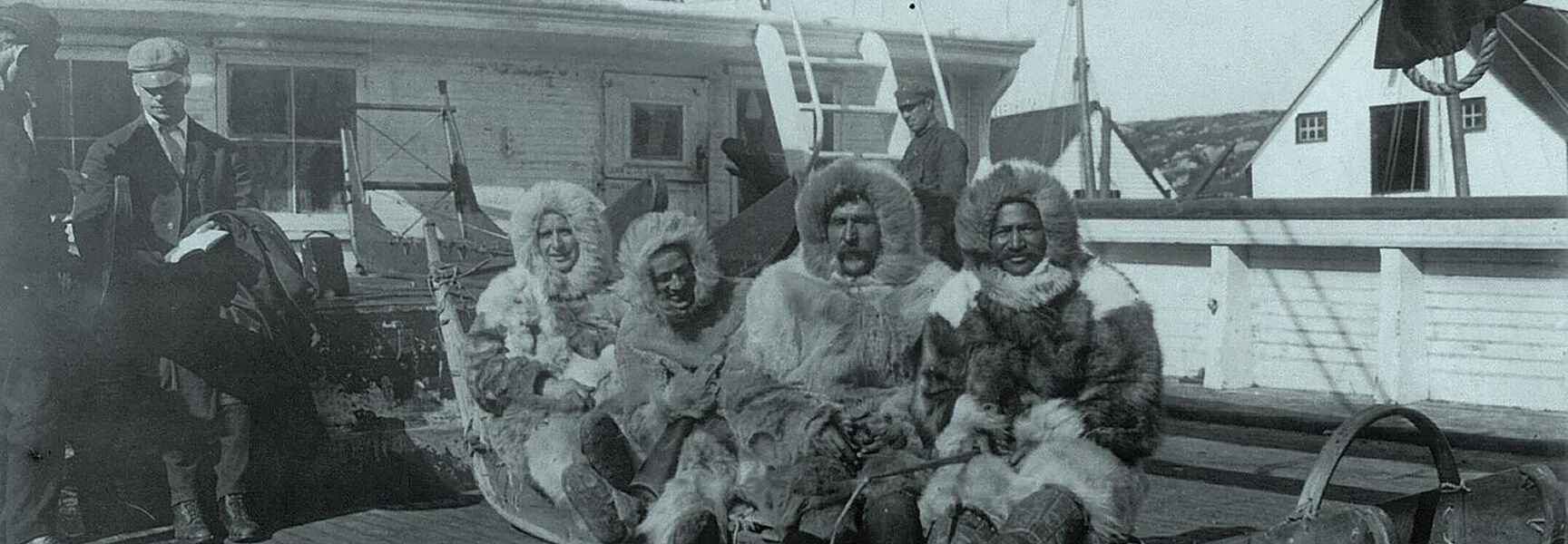 Four men from Peary's North Pole Expedition wearing heavy fur parkas sit on a wooden sled on the deck of a ship.