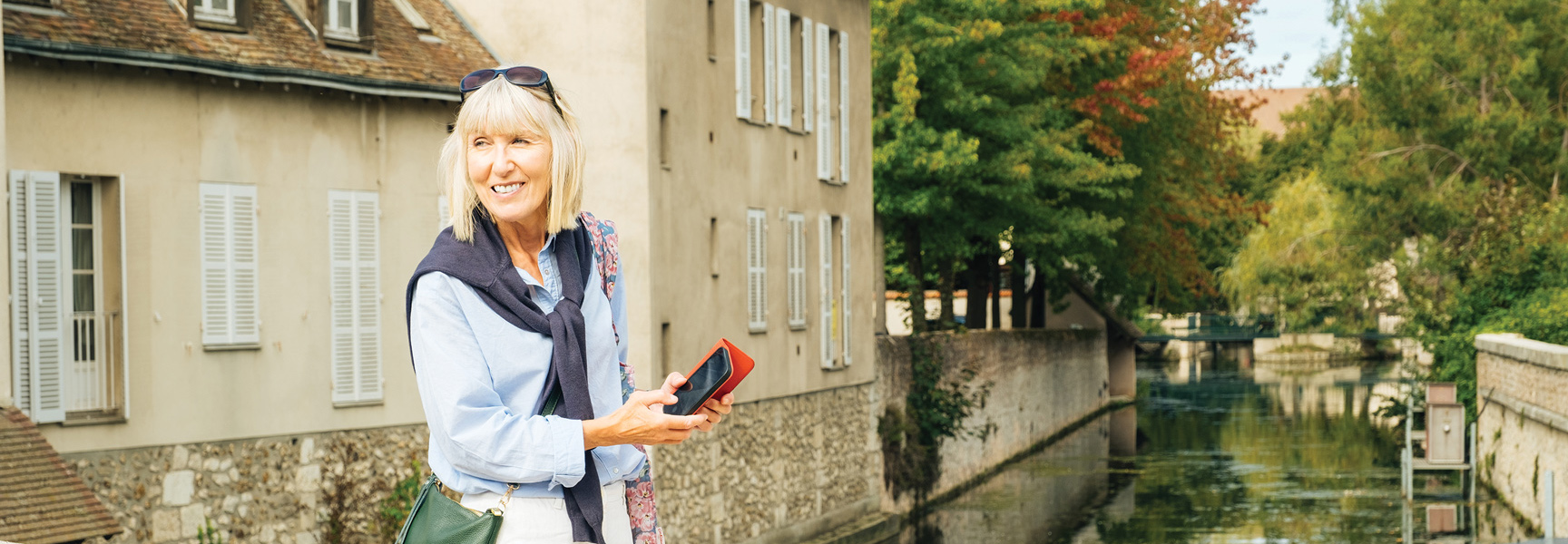 A smiling blonde woman holds a phone while standing by a canal in a picturesque town in France.