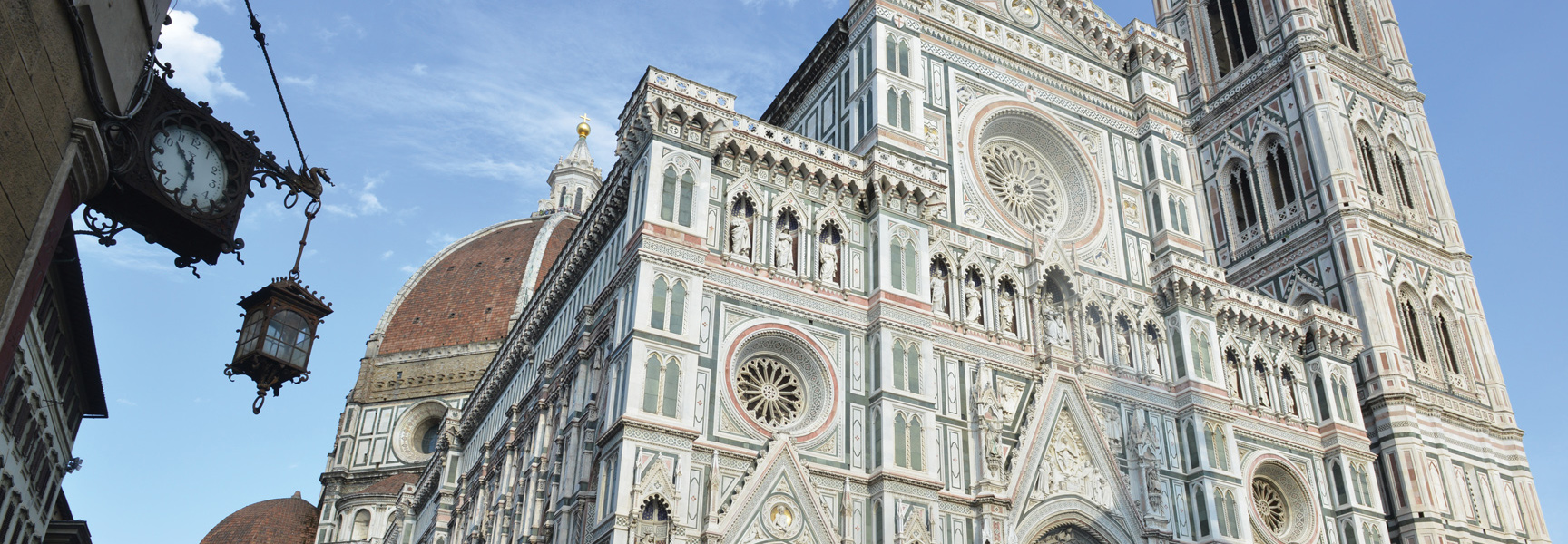 The ornate, detailed marble facade of the Duomo in Florence, Italy, with its famous dome under a bright blue sky.