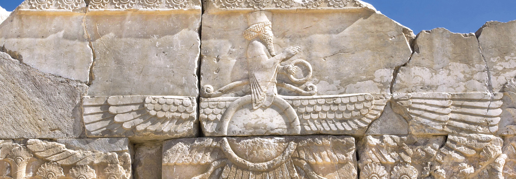 A close-up of an ancient Persian stone carving of a Faravahar, a winged figure, set against a blue sky.