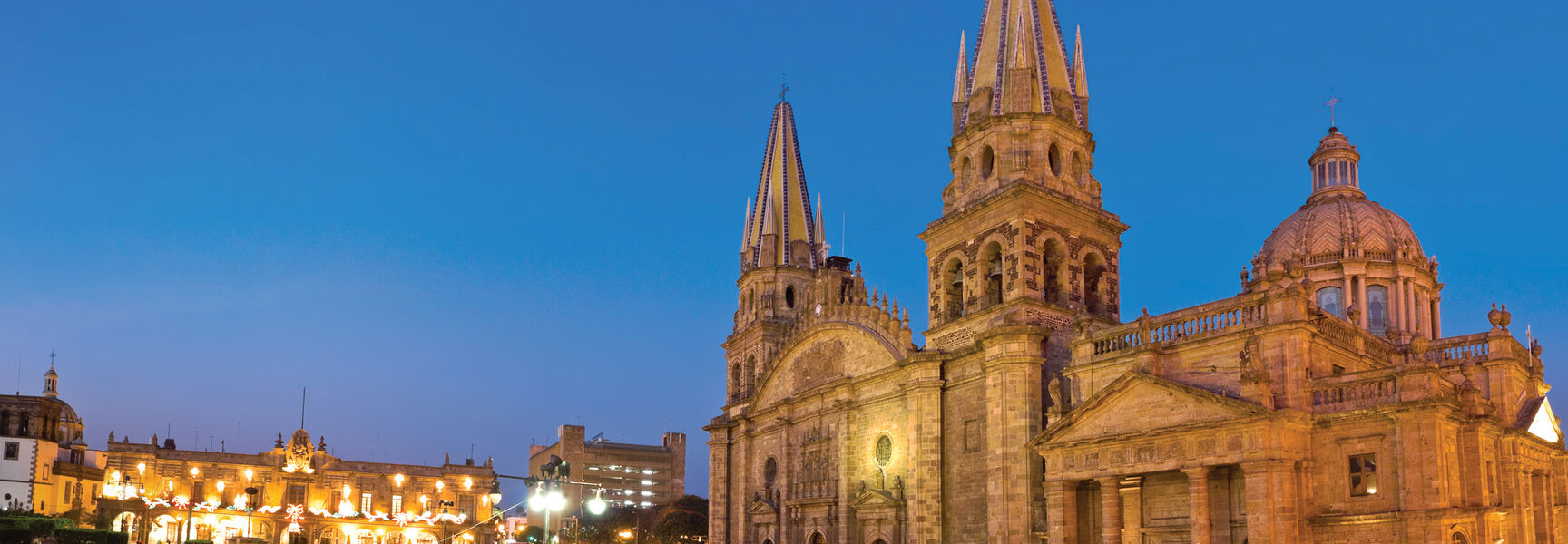 The Guadalajara Cathedral in Mexico at twilight, featuring illuminated twin spires and a bustling city square with long-exposure light trails from passing vehicles.