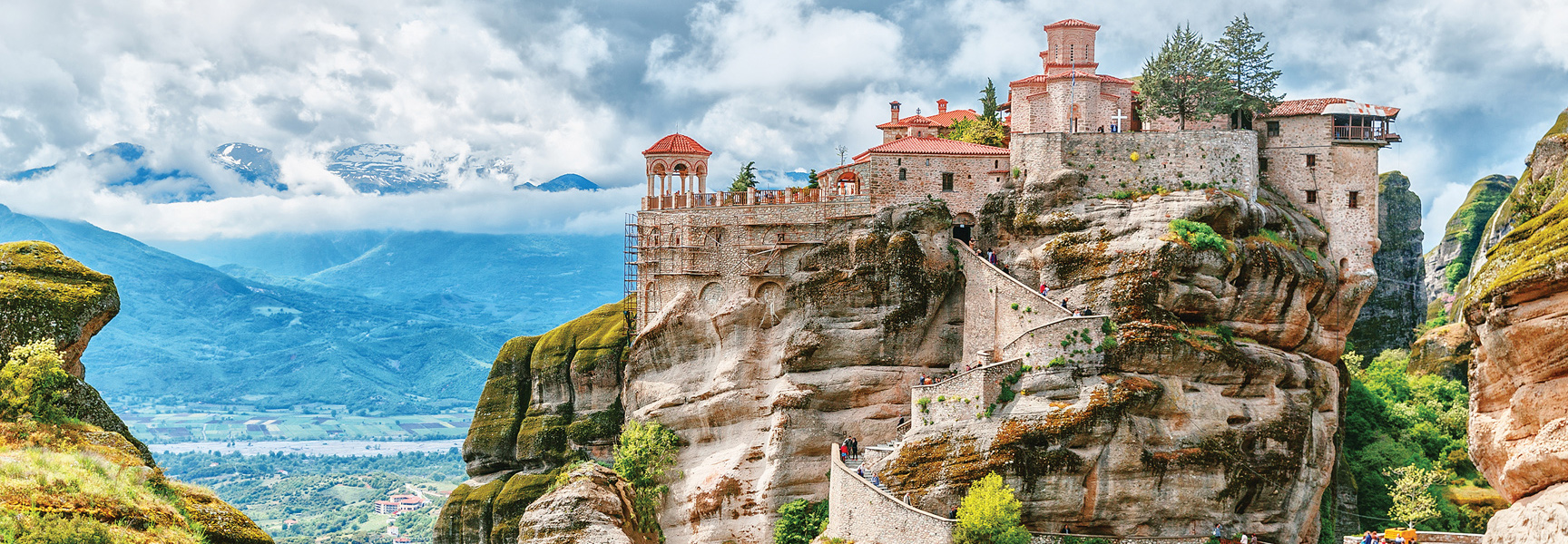 A monastery with red-tiled roofs is built into a dramatic cliffside in Meteora, Greece, overlooking a lush valley and distant mountains.