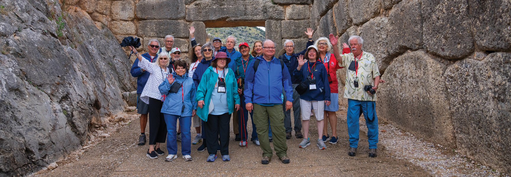 A group of older adult travelers smiles and waves while standing in an ancient stone passageway in Greece.