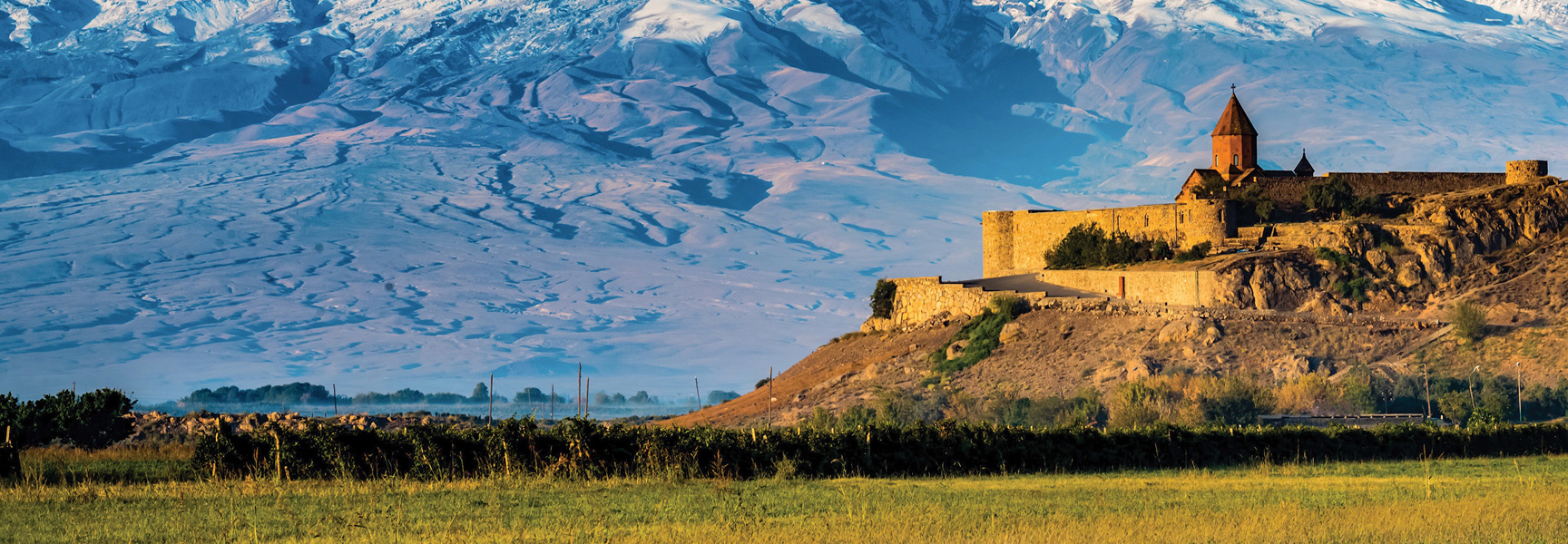A stone monastery on a hill with large, snow-capped mountains in the background and a green field in the foreground.