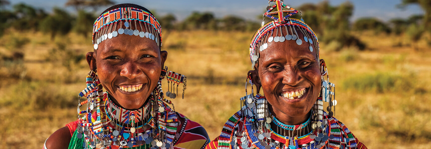 Two smiling women in traditional Kenyan beaded jewelry and colorful clothing stand in a savanna landscape with Mount Kilimanjaro in the background.