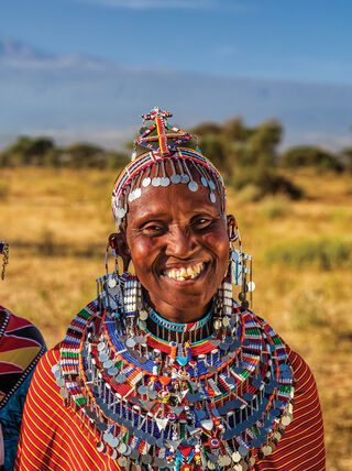 Two smiling women in traditional Kenyan beaded jewelry and colorful clothing stand in a savanna landscape with Mount Kilimanjaro in the background.