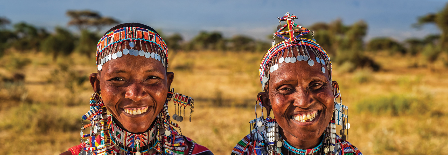 Two smiling Kenyan women in colorful, traditional beaded attire and headdresses stand in the savanna.
