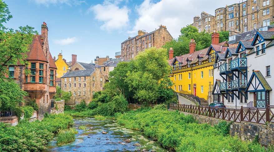 Colorful, historic buildings of Dean Village in Edinburgh, Scotland, line the lush green banks of the Water of Leith under a partly cloudy sky.