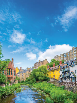 Historic colorful buildings line the Water of Leith in Dean Village, Edinburgh, Scotland, under a vibrant blue sky with scattered clouds.