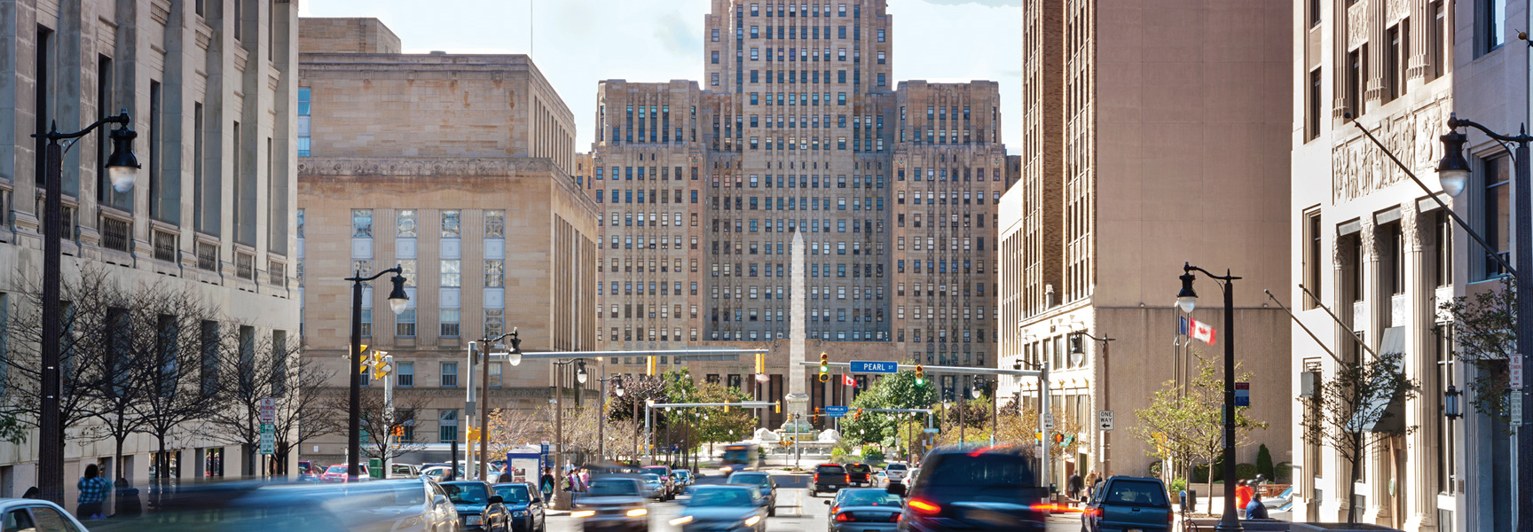 A bustling street-level view of downtown Buffalo, New York, showcases historic architecture and blurred traffic on a bright day.