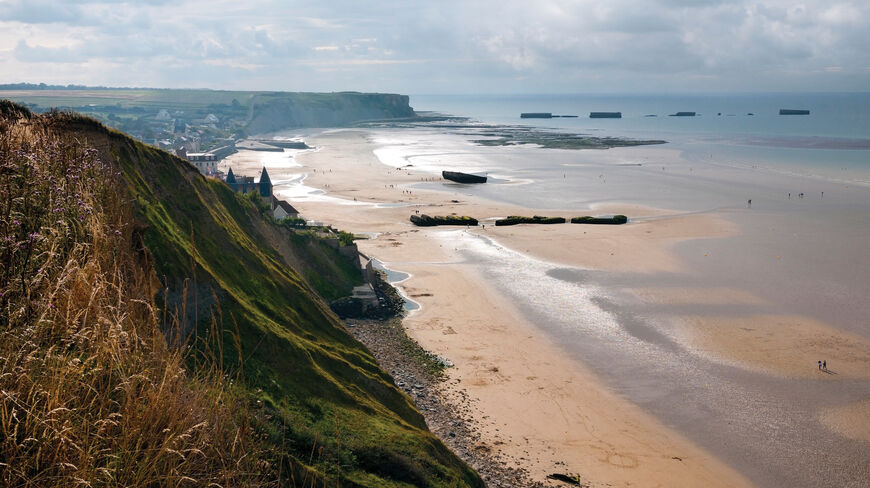A panoramic view of a Normandy beach at low tide featuring the historical remnants of the D-Day Mulberry harbor and the surrounding coastal cliffs.