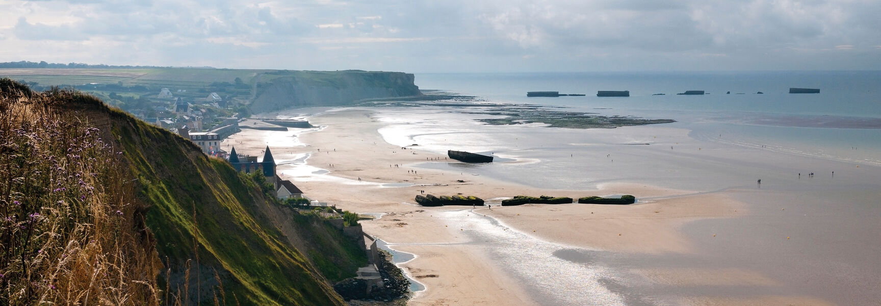 A panoramic view of a Normandy beach at low tide featuring the historical remnants of the D-Day Mulberry harbor and the surrounding coastal cliffs.
