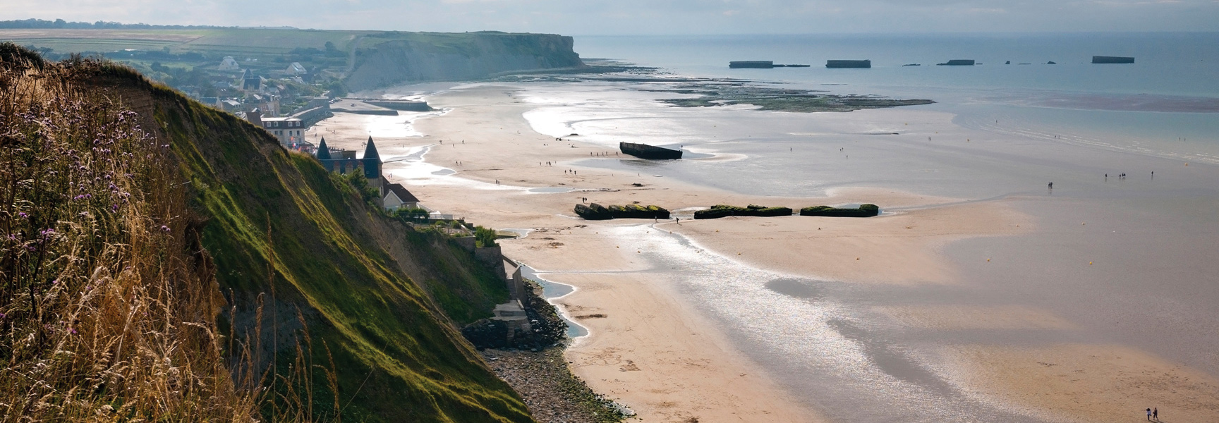 The historic D-Day beaches at Arromanches, Normandy, with remnants of the Mulberry harbor on the sand and in the sea at low tide.