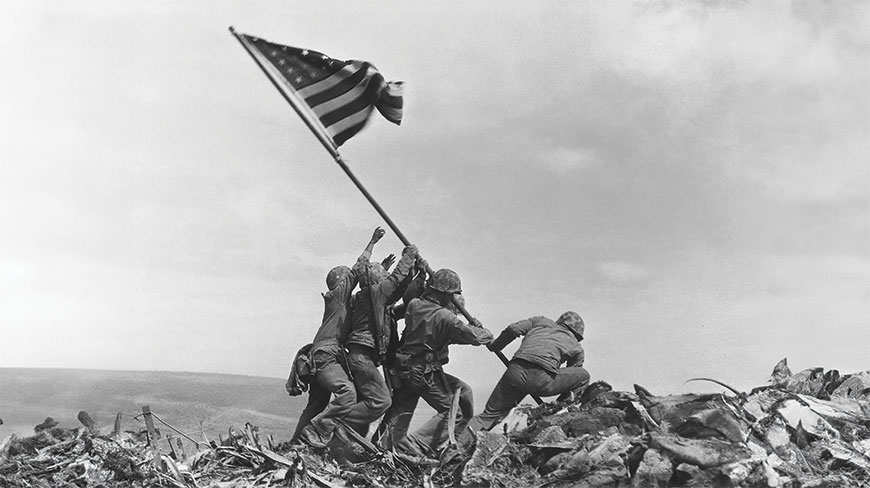 A black and white photograph of several soldiers raising an American flag on a pole atop a rocky hill.