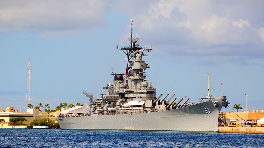 The historic Battleship USS Missouri is docked in the blue waters of Pearl Harbor on a partly cloudy day.