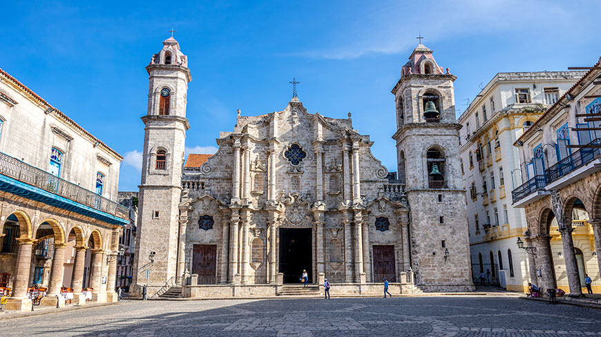 The Havana Cathedral, an ornate stone building with two towers, stands in a wide, sunny plaza in Cuba.