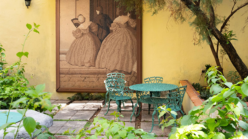 A green wrought-iron table and chairs sit in a sunny courtyard in Havana, Cuba, in front of a large painting on a yellow wall.