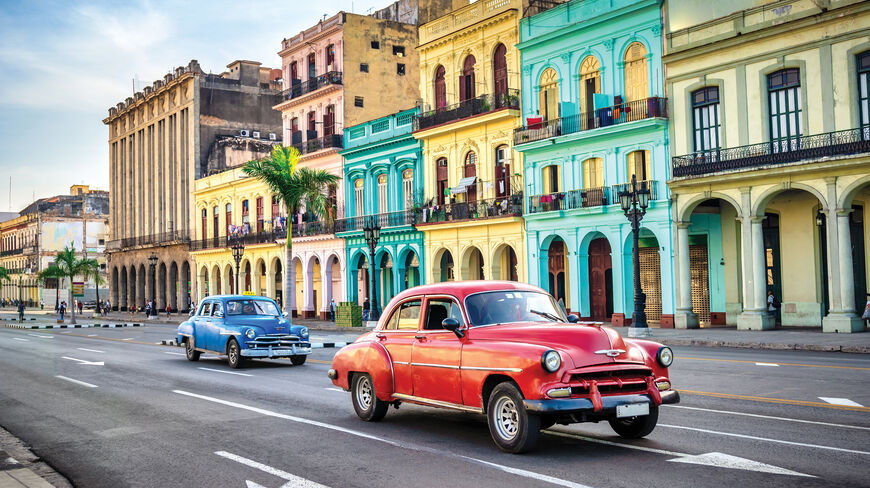 Vintage cars drive past vibrant, colorful colonial architecture on a street in Havana, Cuba.