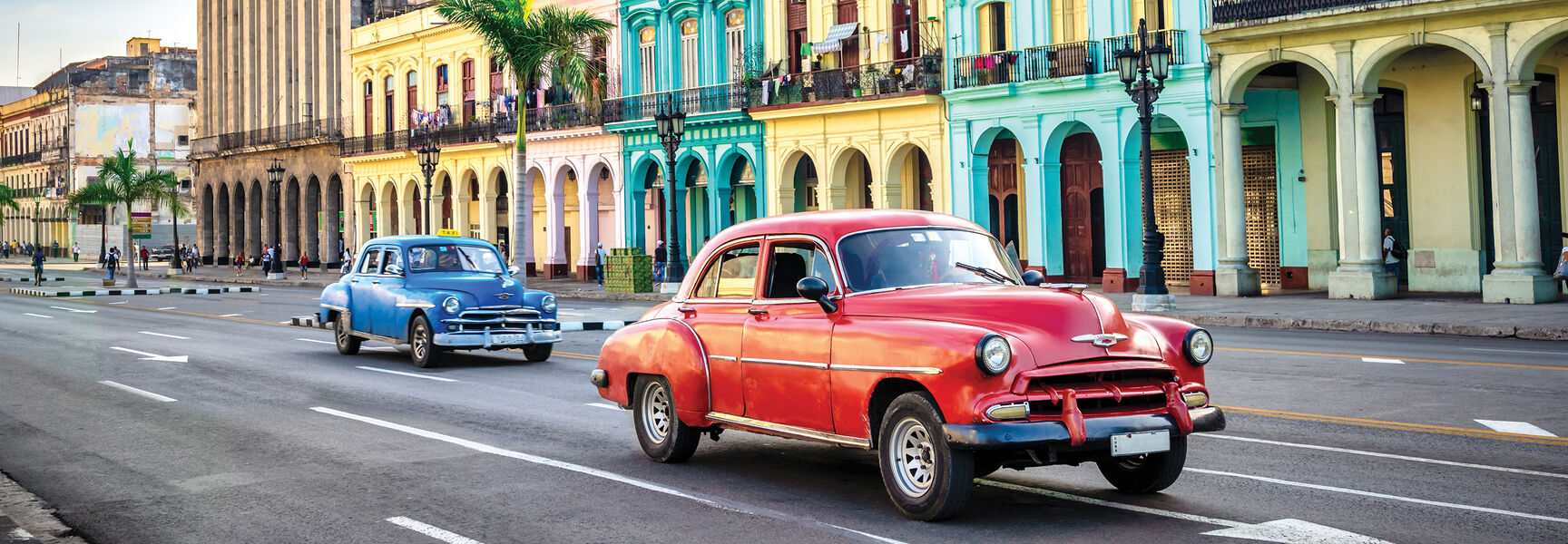 Vintage cars drive past vibrant, colorful colonial architecture on a street in Havana, Cuba.