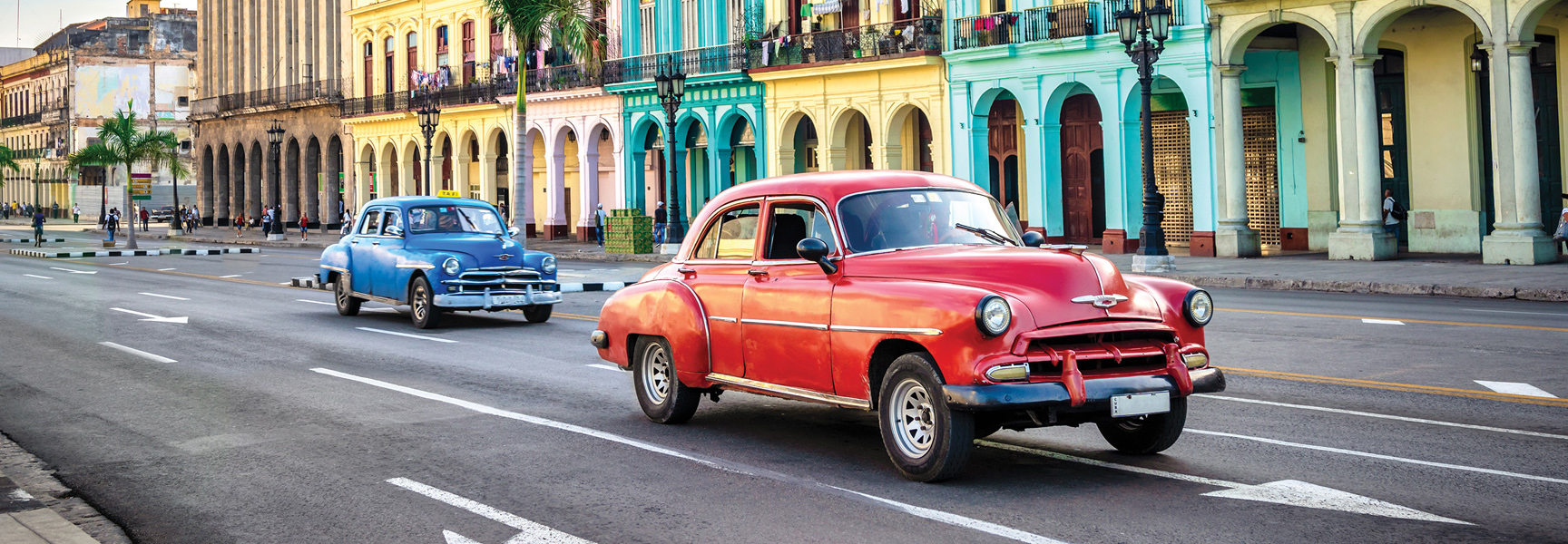 Two classic American cars, one red and one blue, drive past colorful colonial-style buildings on a sunny street in Cuba.