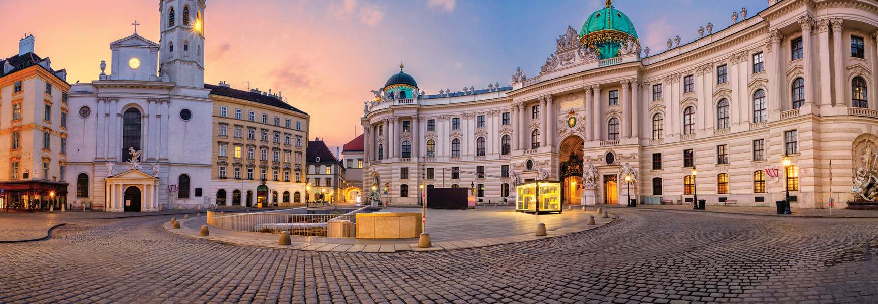 A cobblestone square in Central Europe curves past ornate, historic buildings, including a church and a domed palace, all illuminated at sunset.