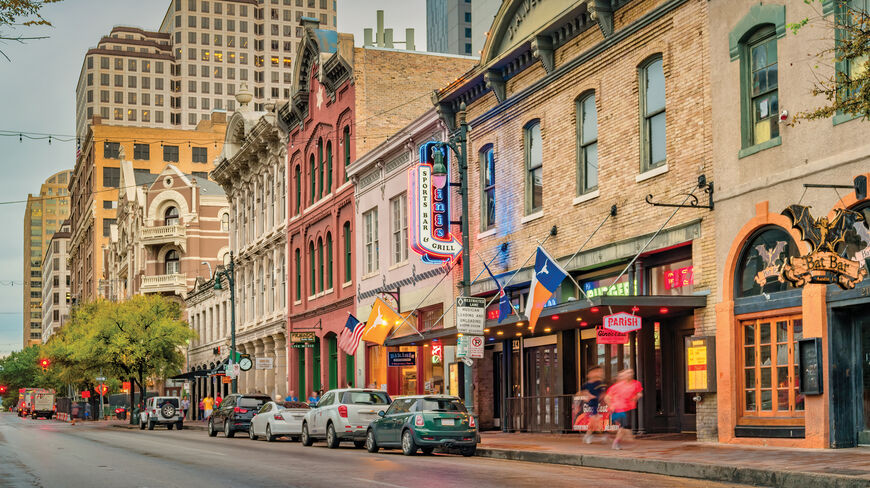 A street view of the historic district in Austin, Texas, featuring colorful heritage buildings, storefront signs, and flags along the sidewalk at dusk.