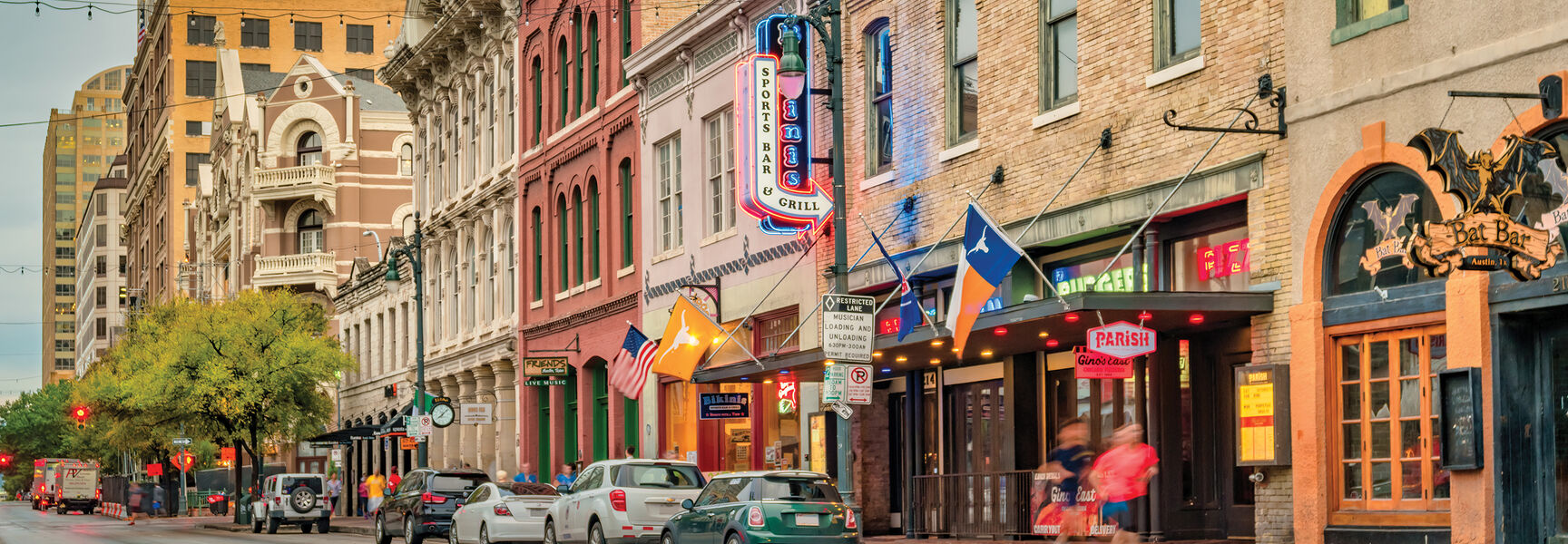 A street view of the historic district in Austin, Texas, featuring colorful heritage buildings, storefront signs, and flags along the sidewalk at dusk.