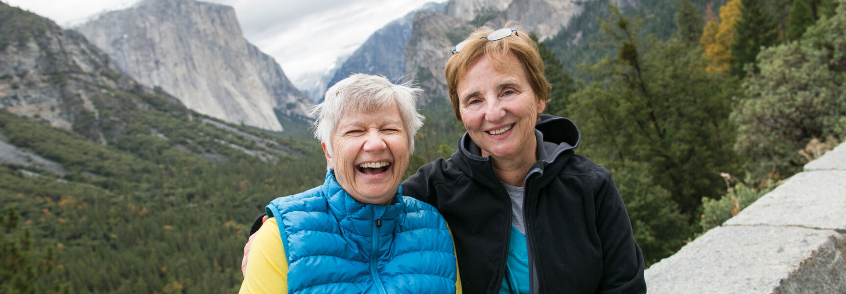Two women laughing on a hiking adventure in Yosemite National Park, California, with scenic views of mountains and glacial valleys in the background.