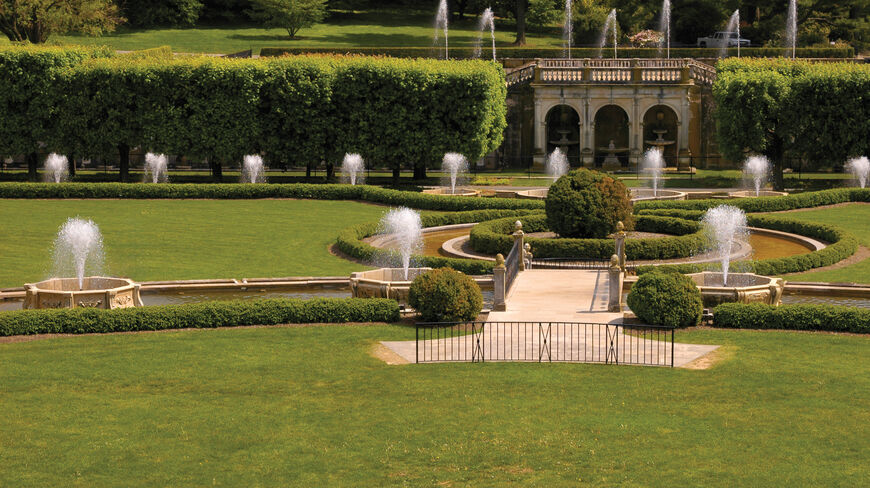 Fountains spray water throughout the manicured botanical pleasure gardens of Pennsylvania, featuring sculpted hedges and a classic stone arched structure.