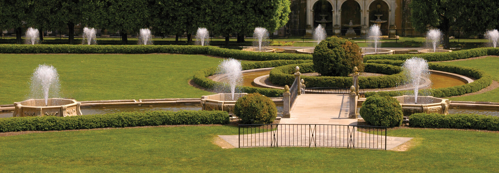 Fountains spray water throughout the manicured botanical pleasure gardens of Pennsylvania, featuring sculpted hedges and a classic stone arched structure.