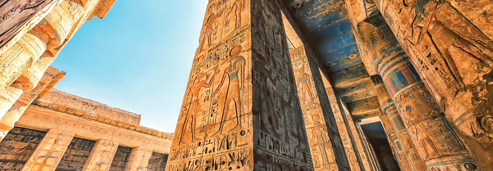 Looking up at large stone pillars covered with colorful hieroglyphics inside an ancient temple in Egypt under a clear blue sky.