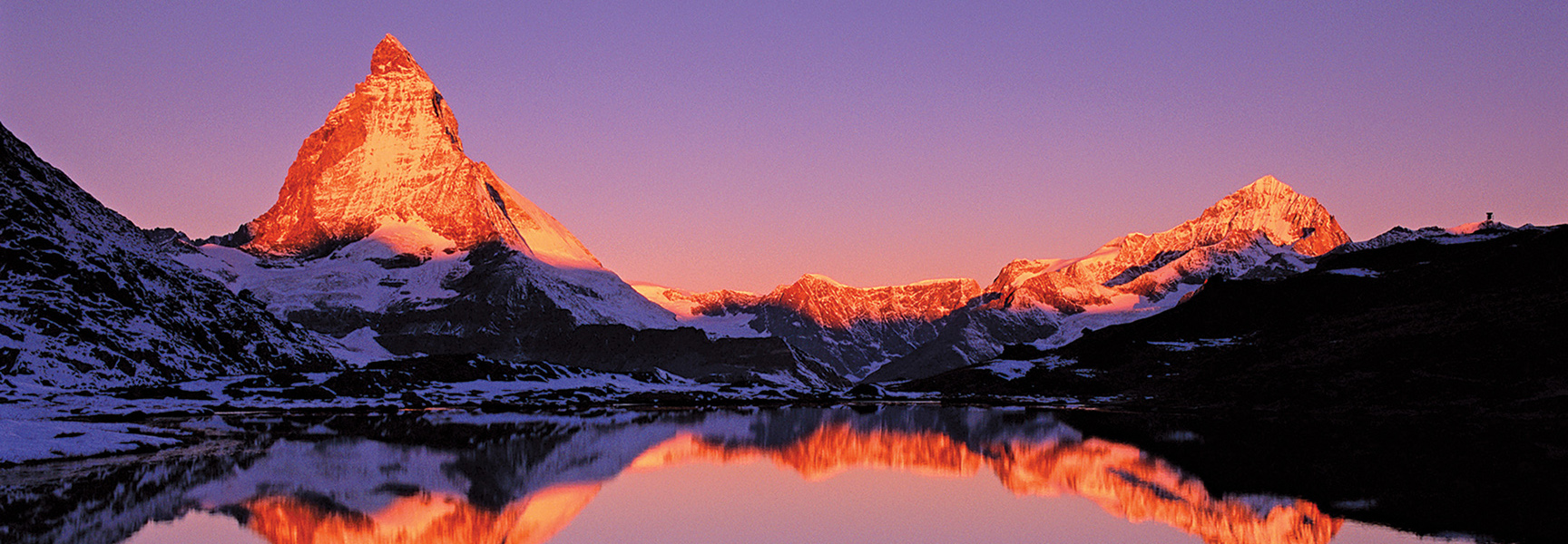 The Matterhorn mountain in Switzerland is reflected in a lake as the rising sun illuminates its peak against a purple sky.