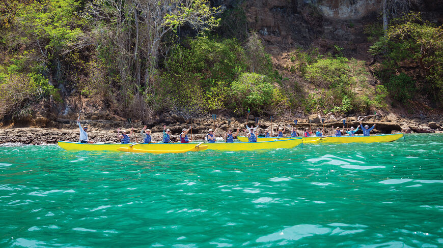 A group of solo travelers in yellow outrigger boats paddle across turquoise water near the rocky coastline of Costa Rica.