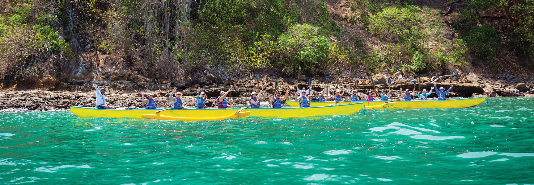 A group of solo travelers in yellow outrigger boats paddle across turquoise water near the rocky coastline of Costa Rica.