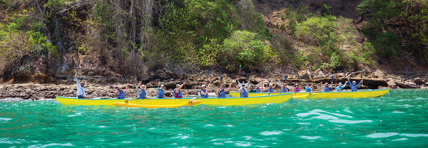 A group of travelers paddle bright yellow outrigger canoes on turquoise water along the lush, rocky coast of Costa Rica.