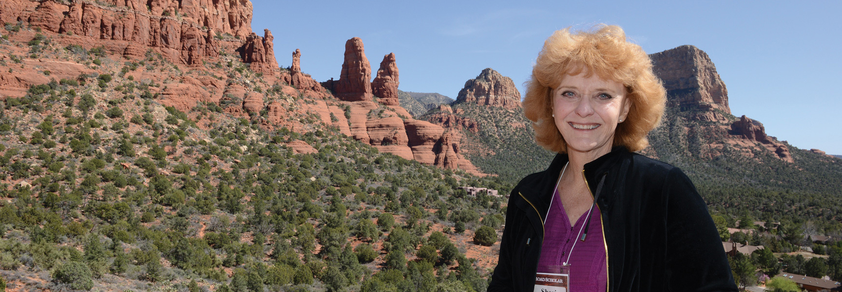A woman smiles in front of the beautiful red rock formations of Sedona, Arizona on a sunny day.