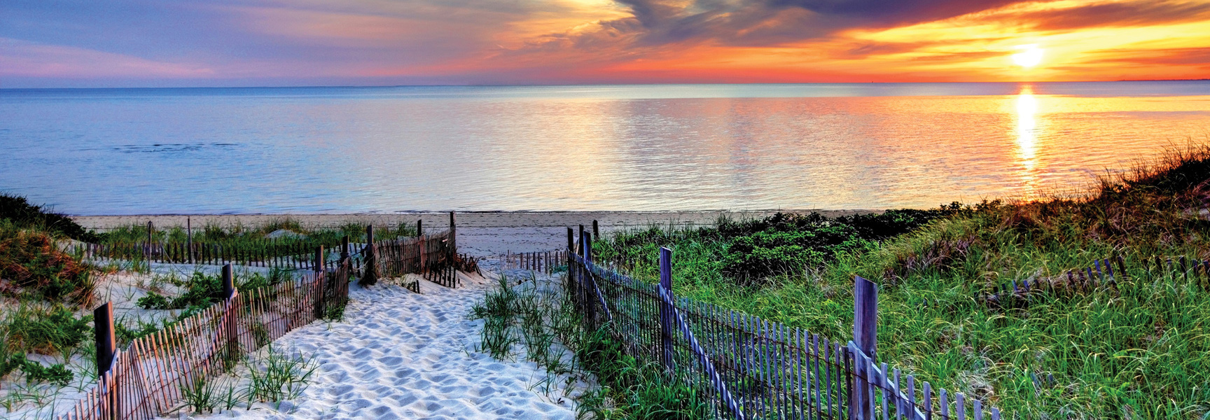A sandy path through grassy dunes leads to a calm ocean at sunset on Cape Cod, Massachusetts.