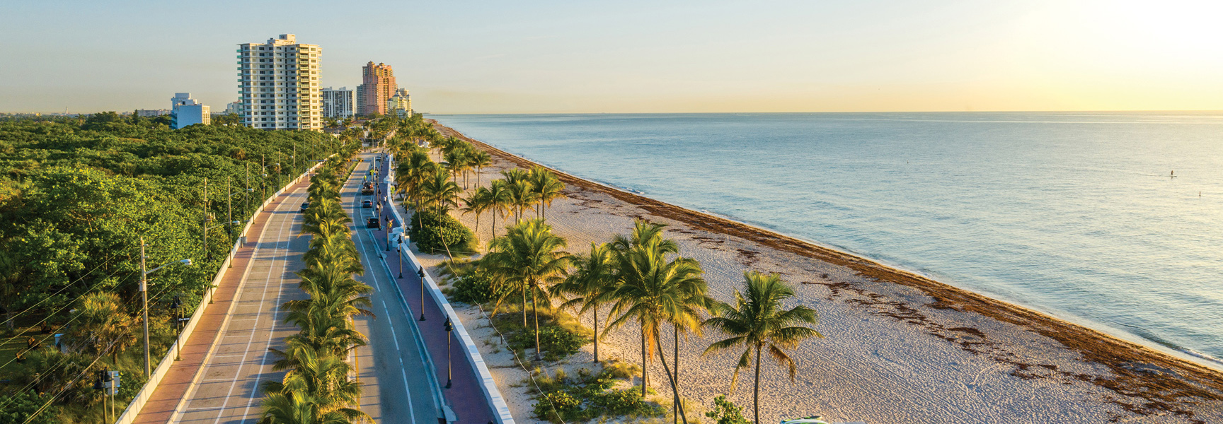 An aerial view of a scenic highway running alongside a sun-drenched Miami beach with palm trees, high-rise buildings, and the ocean at sunrise.