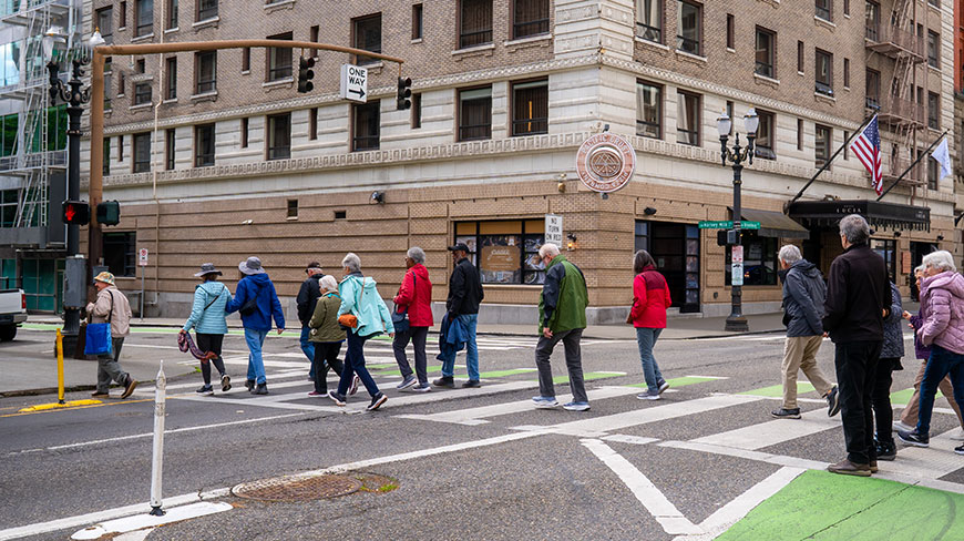 A group of travelers walks across a city street intersection in Portland, Oregon, with historic brick buildings in the background.