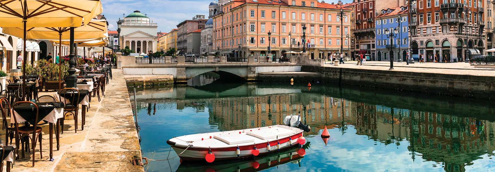 A small boat floats in a canal in Trieste, Italy, lined with outdoor cafes and colorful historic buildings on a sunny day.