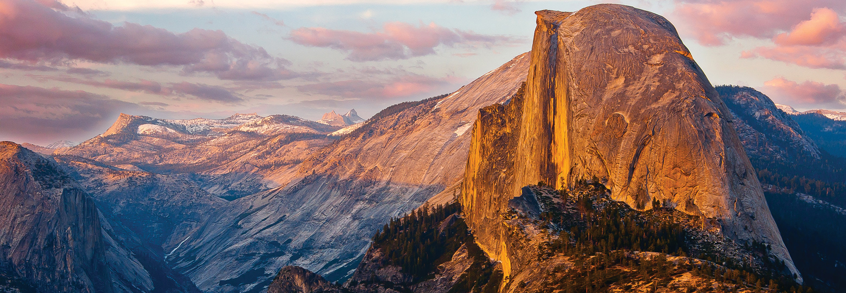 The iconic Half Dome glows with golden sunset light over the vast mountain landscape of Yosemite National Park in California.