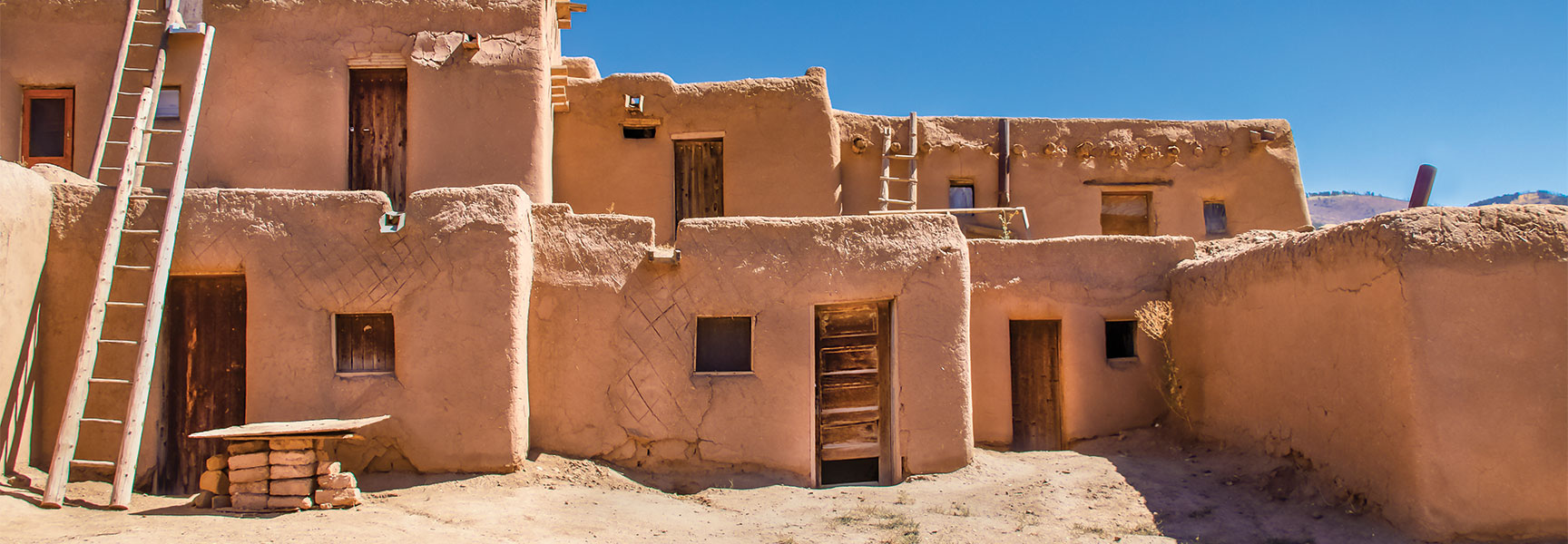 Multi-storied adobe Pueblo houses in New Mexico featuring wooden ladders and doors against a bright blue sky.