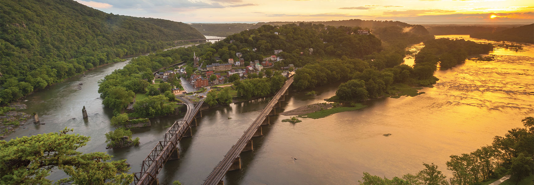 Aerial sunset view of Harpers Ferry with bridges crossing the confluence of the Potomac and Shenandoah rivers, surrounded by lush green mountains.