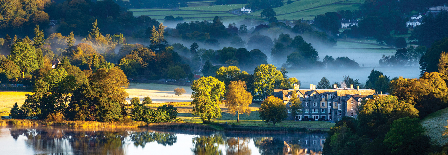A large stone hotel on a lake is surrounded by rolling green hills and trees with a light mist in England's Lake District.