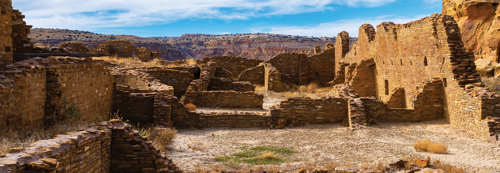 The ruins of ancient stone buildings sit within a desert canyon under a partly cloudy blue sky.